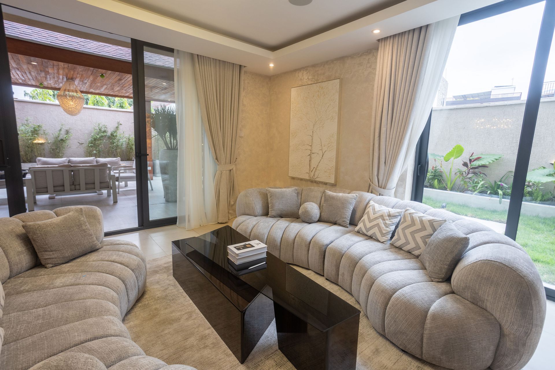 Modern living room with curved beige sofas, glass coffee table, and large windows overlooking a patio.