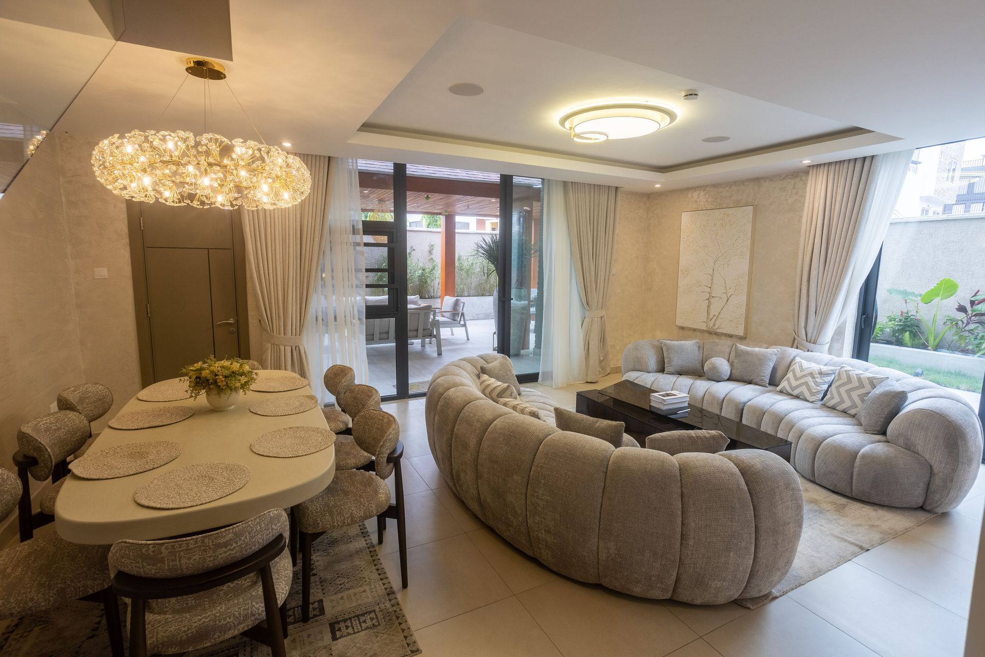 Elegant beige living room with plush curved sofas, chandelier, dining table, and glass doors to a patio