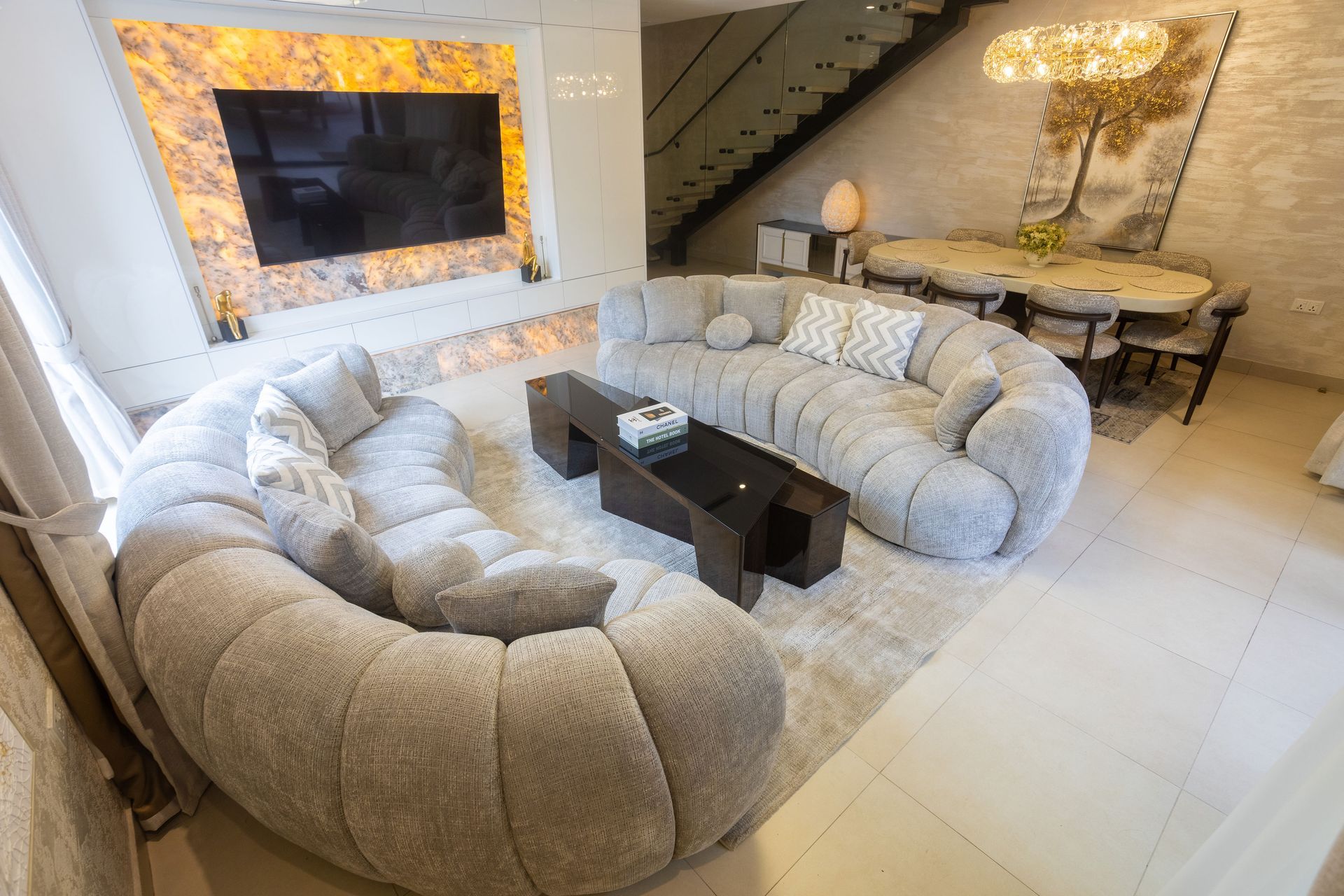 Modern living room with curved gray sofas, a wall-mounted TV, and a glass dining area with warm lighting