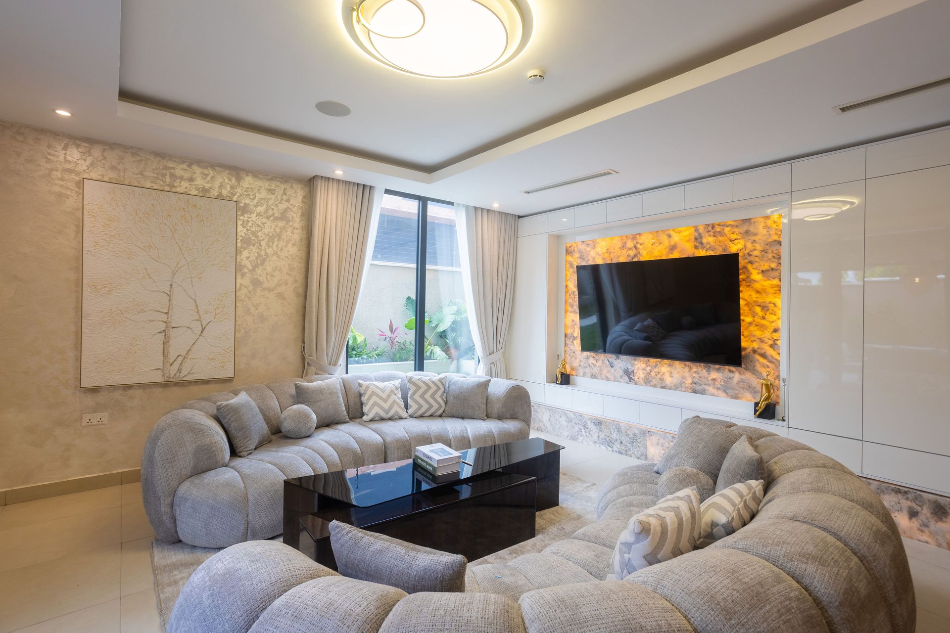 Bright modern living room with curved gray sofas, black coffee table, and wall-mounted TV above a fireplace.