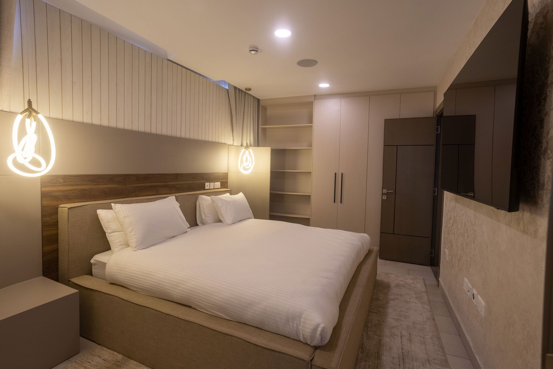 Modern beige bedroom with a large bed, pendant lights, built-in shelves, and a wall-mounted TV