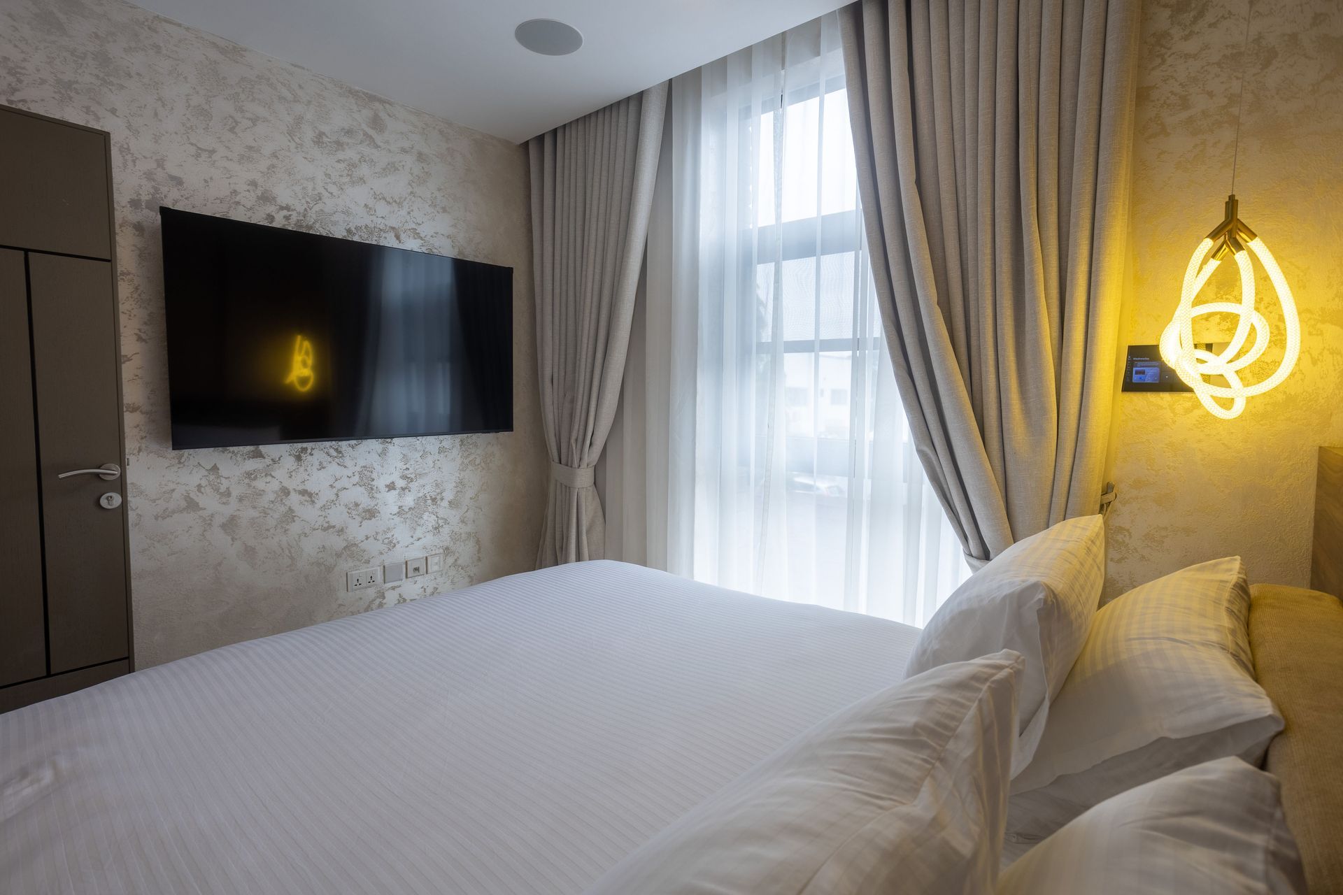 Modern hotel bedroom with white bed, wall-mounted TV, sheer curtains, and warm bedside light
