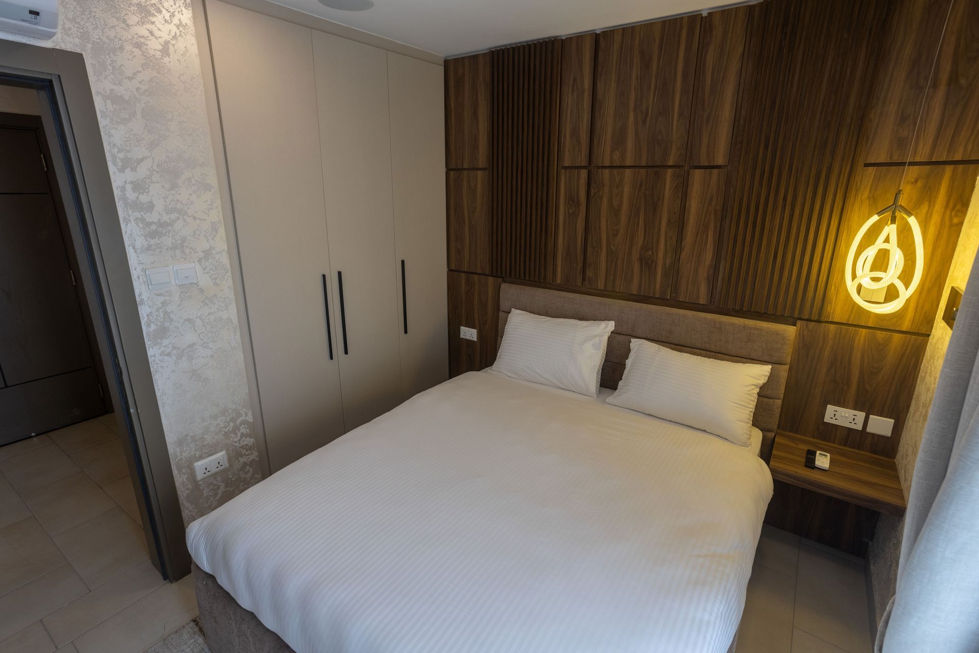 Modern hotel bedroom with a double bed, wood-paneled headboard, wardrobe, and wall lamp