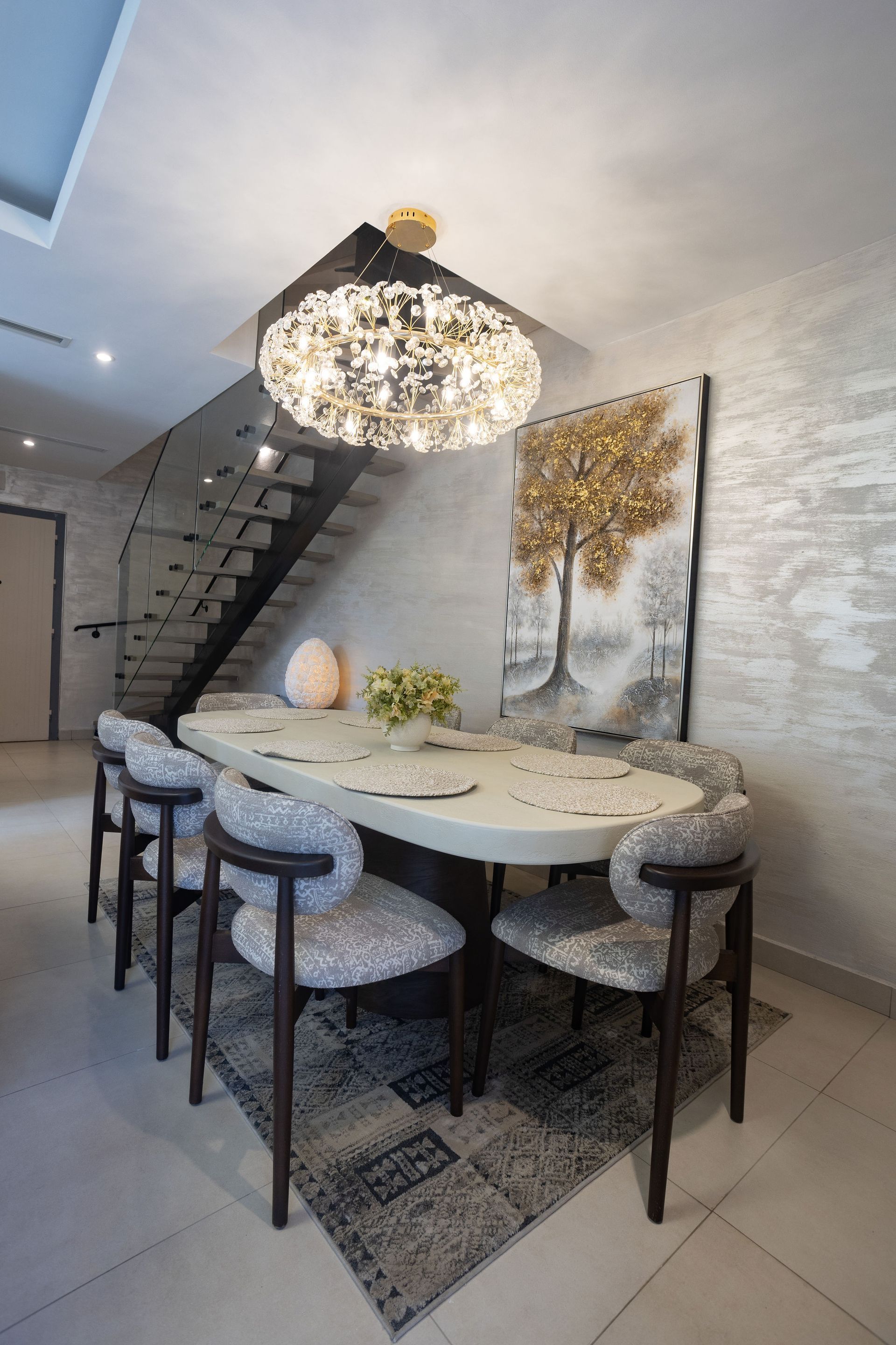 Modern dining room with round table, patterned chairs, chandelier, and staircase.
