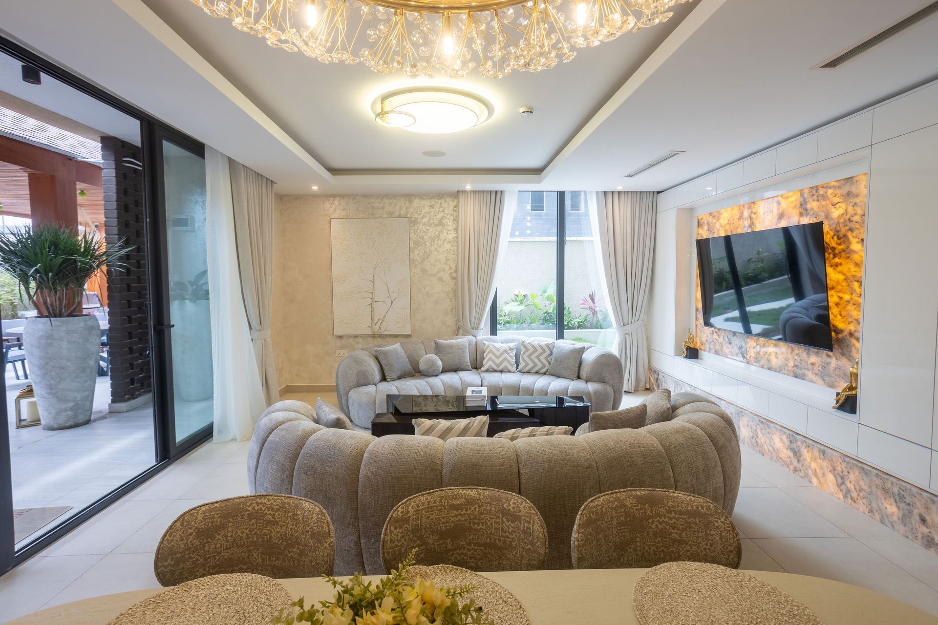 Modern living room with gray sofas, chandelier, large windows, and a wall-mounted TV