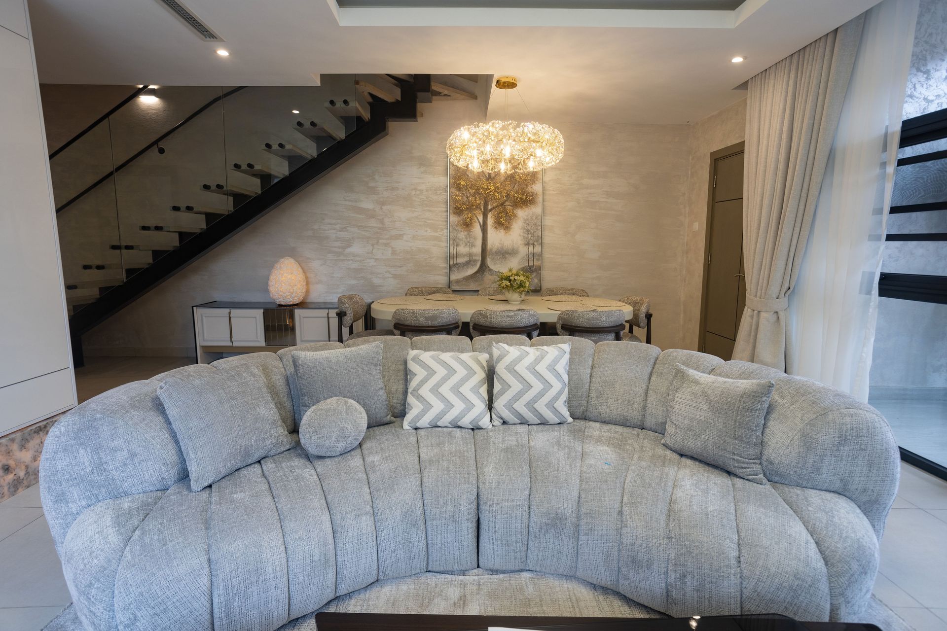 Elegant living room with curved gray sofa, chandelier, staircase, and console table with flowers