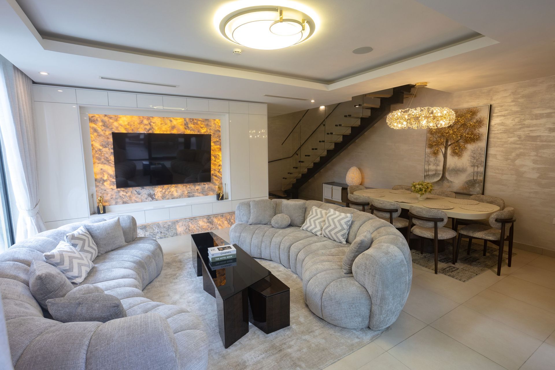 Modern living room with curved cream sofas, wall-mounted TV, and staircase beside a dining area