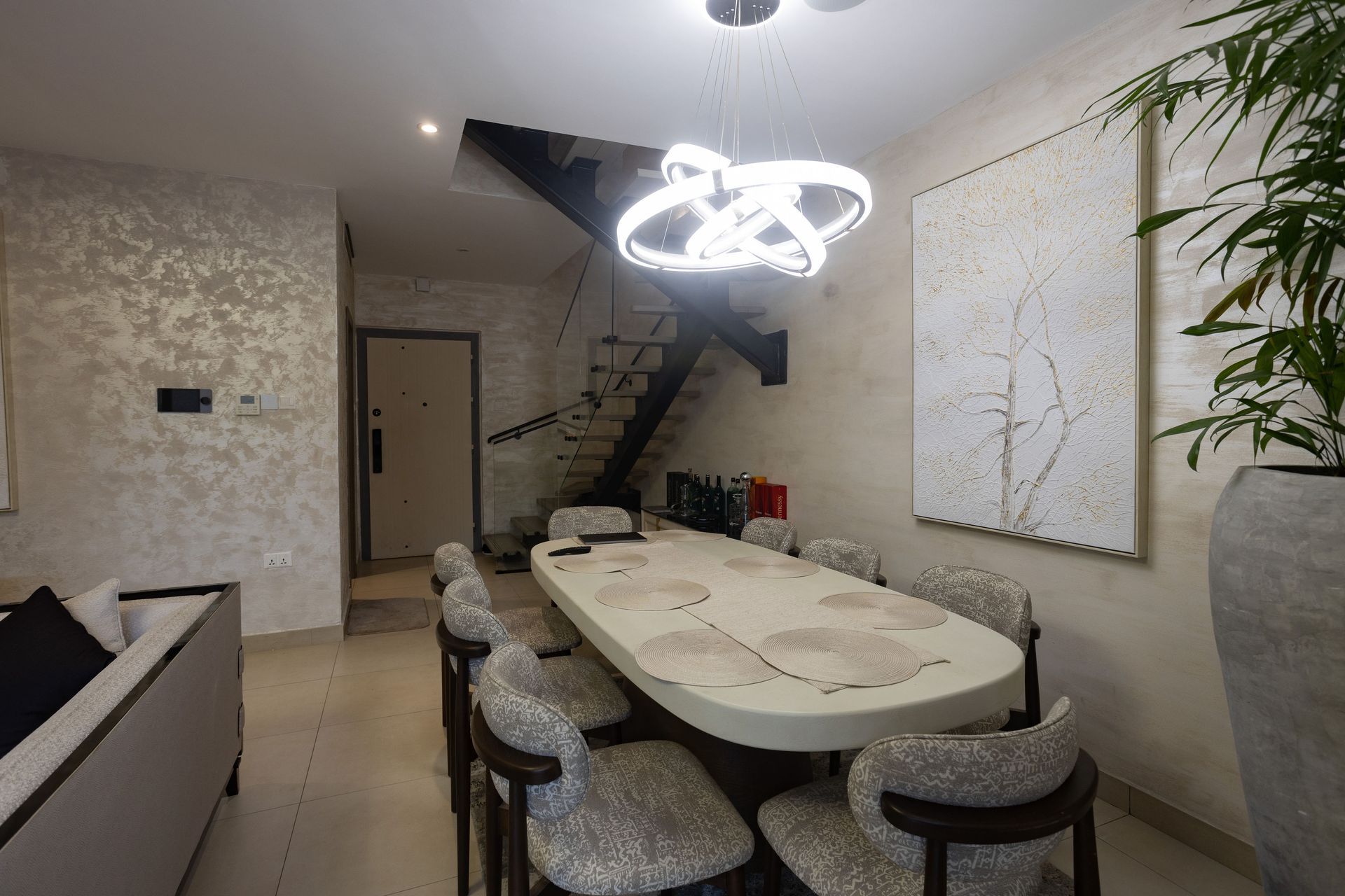 Modern dining room with oval table, patterned chairs, wall art, staircase, and glowing chandelier.