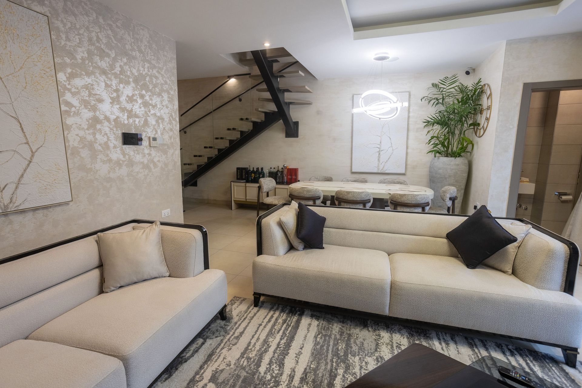 Bright living room with beige sofas, patterned rug, staircase, and potted plant.