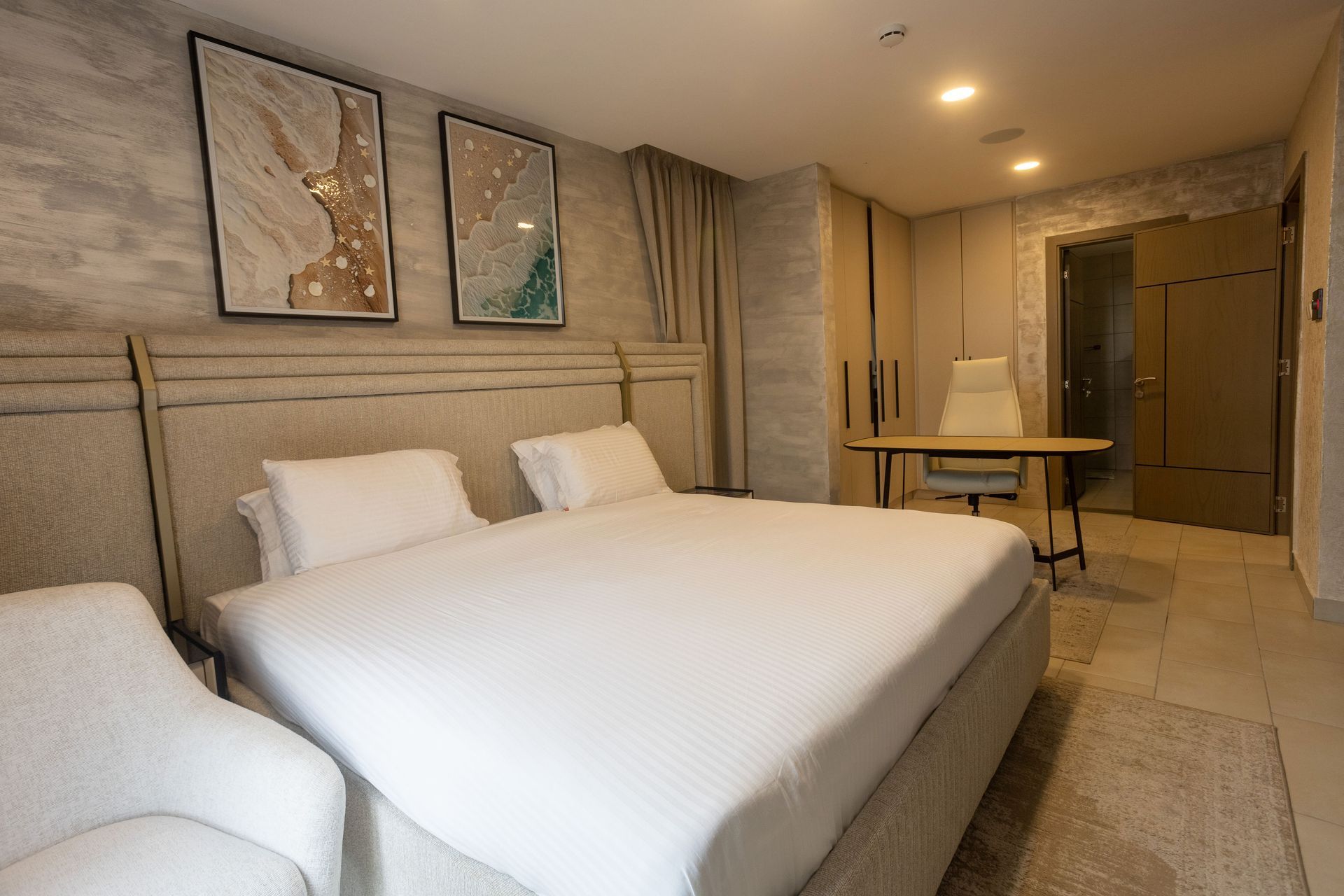 Modern beige bedroom with large bed, wall art, desk, and warm recessed lighting