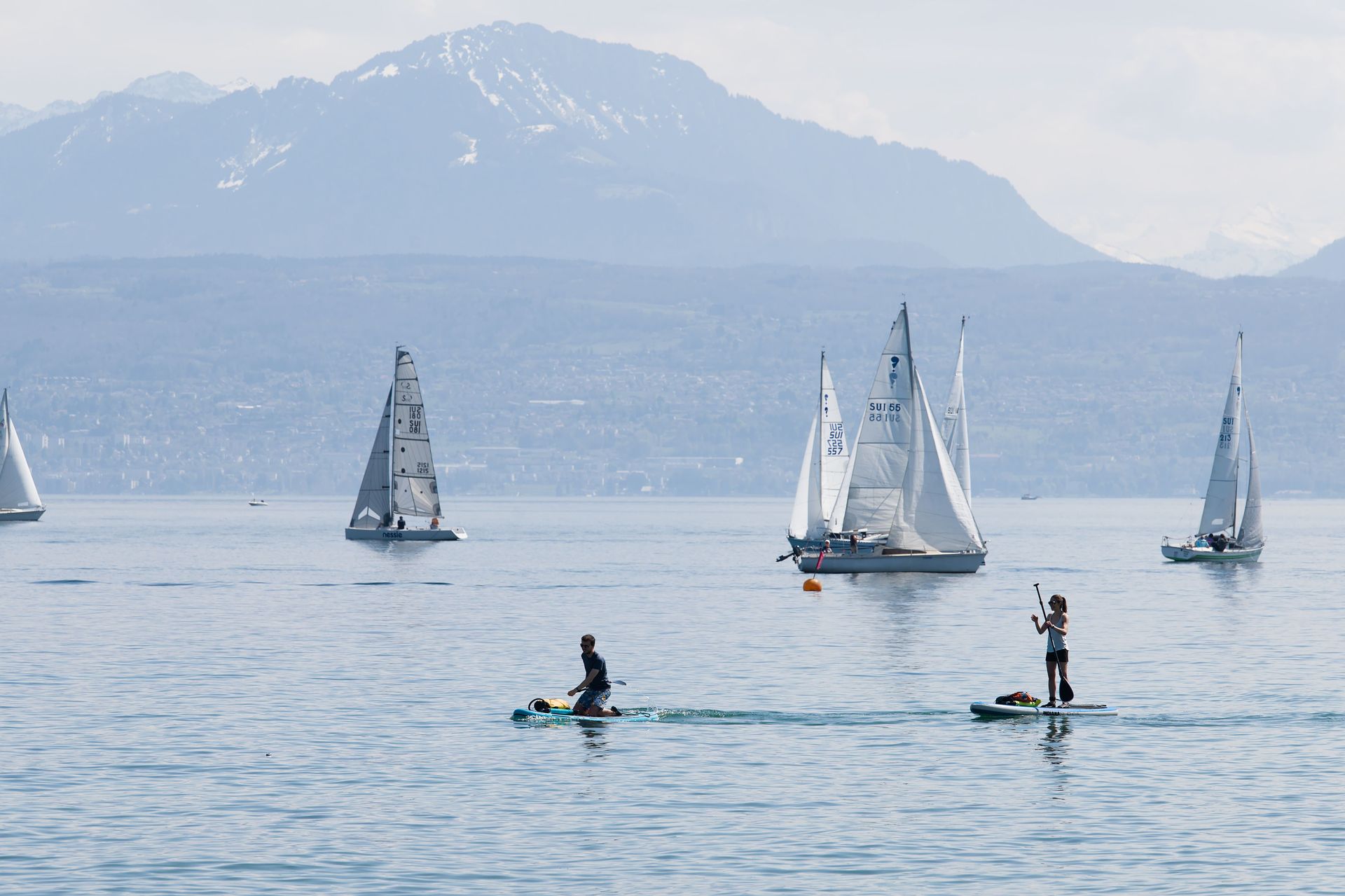 Sailboats on calm water with two people paddleboarding, mountains in the hazy background