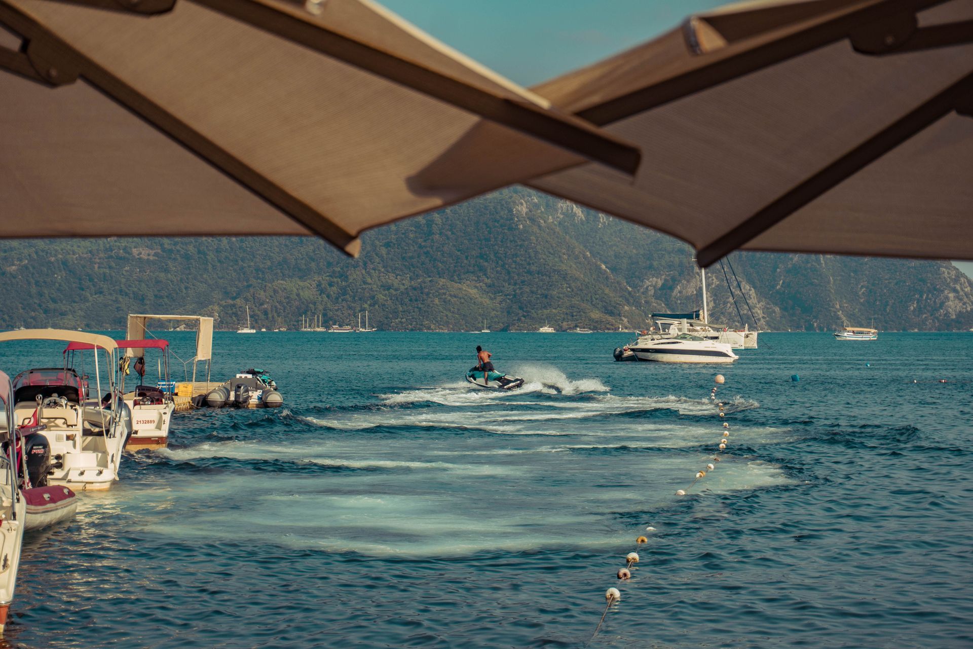 Seaside view from a shaded patio, with boats on turquoise water and mountains in the distance
