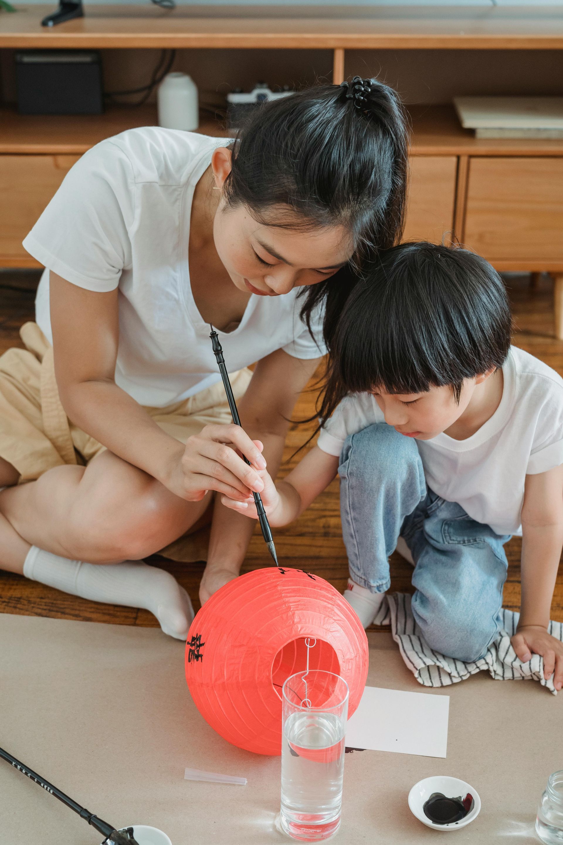 Two children painting a red paper lantern on the floor beside a glass of water.