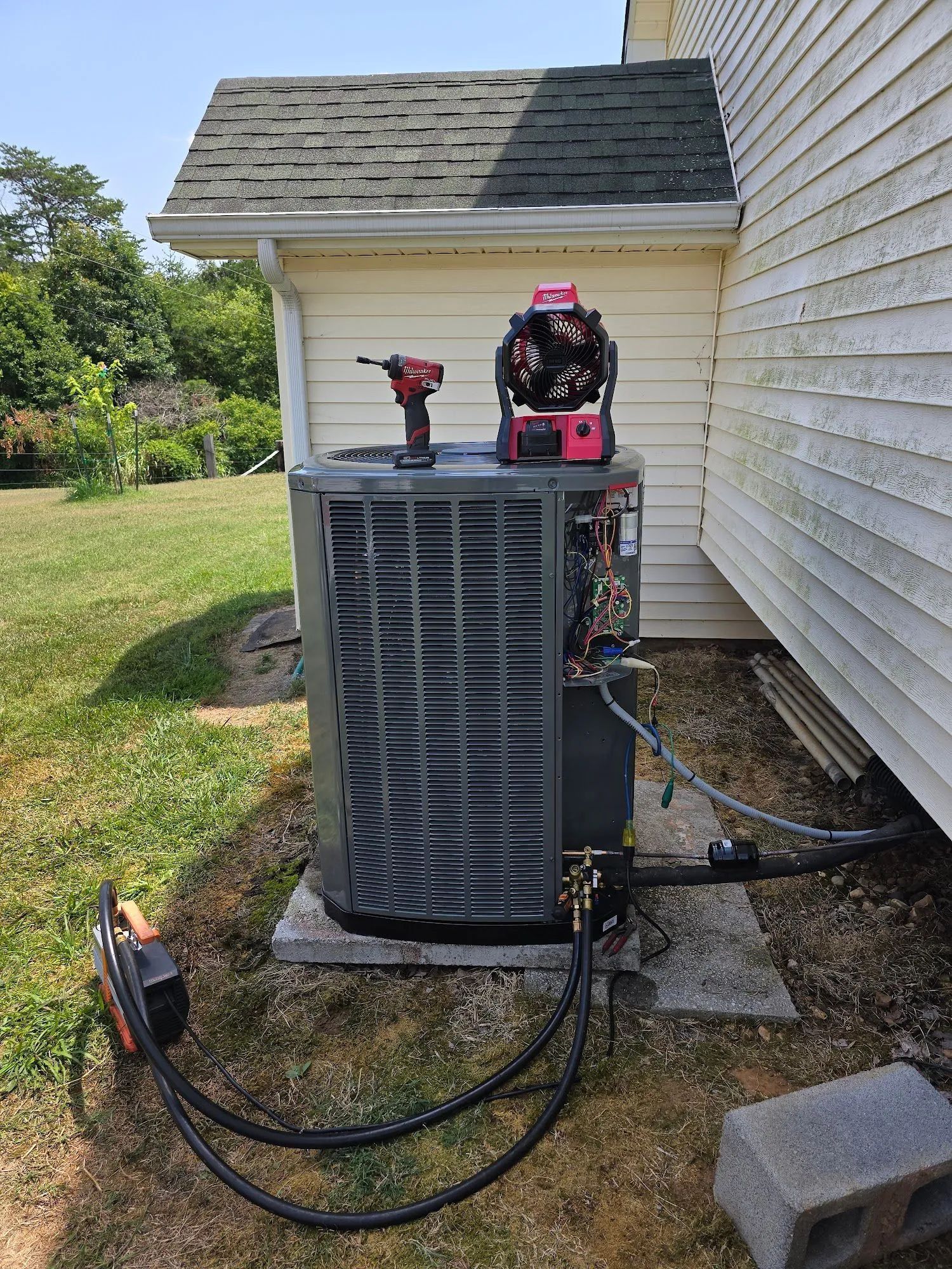Outdoor HVAC unit with a red drill and pink tool bag on top, hoses attached beside a house.