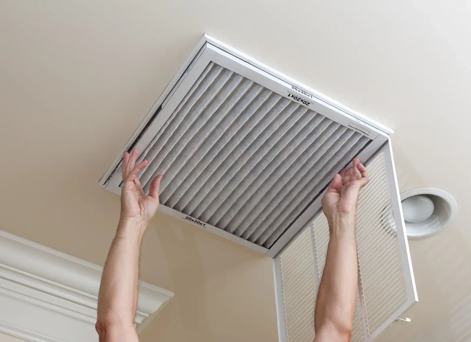Hands removing a ceiling vent cover with a pleated filter from a white ceiling