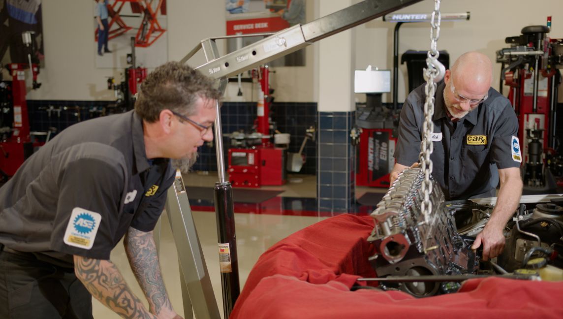 Two mechanics in a shop lift an engine with a hoist. One holds a panel.