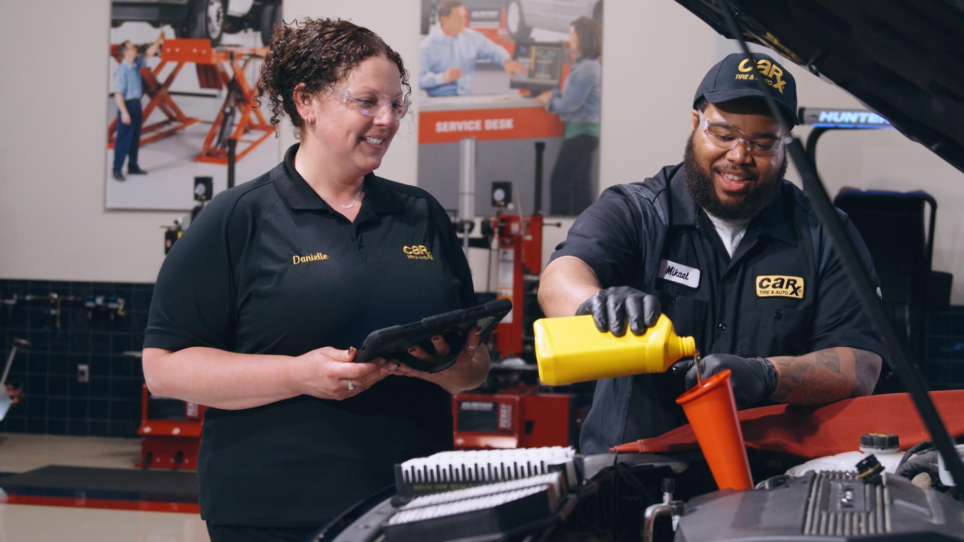 Woman and man in auto shop changing oil; smiling, wearing uniforms.