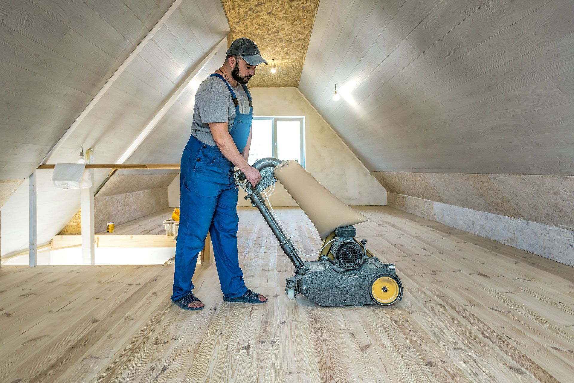 Man sanding wooden floor with a large machine in an attic with slanted walls.