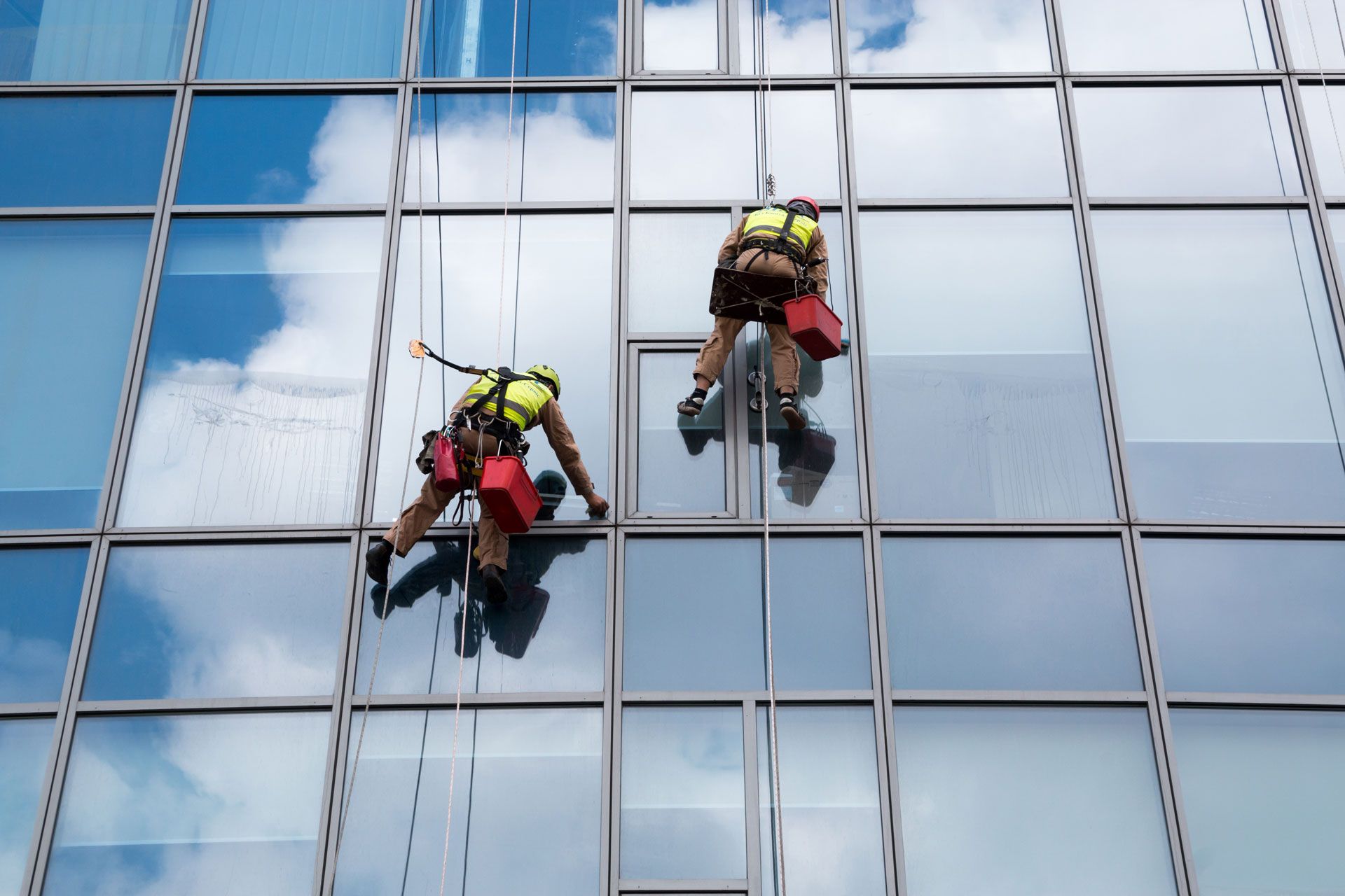 Two window washers on ropes cleaning a glass building with the sky reflected.
