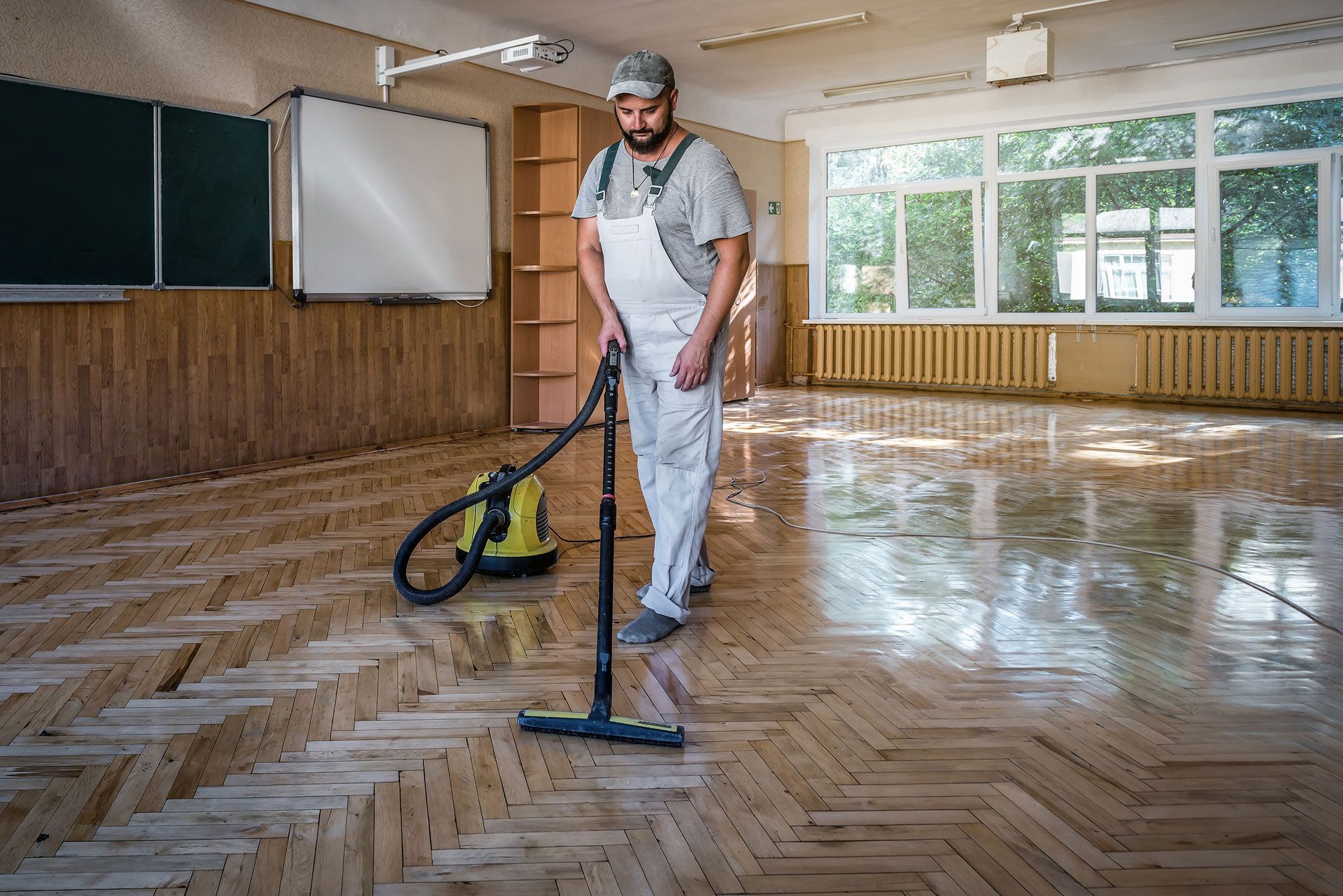 Person in white overalls vacuums a wooden floor in a room with a large window and blackboard.
