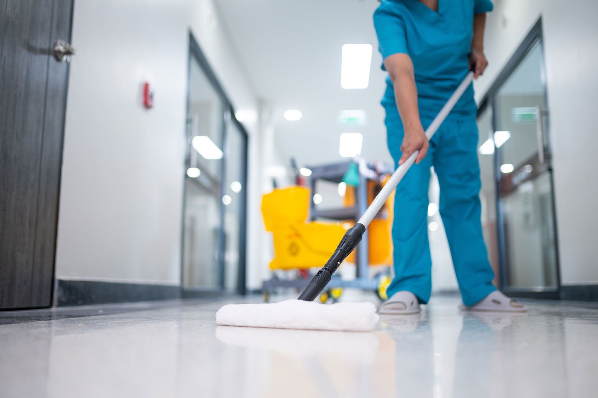 Person in blue scrubs mopping a shiny hallway floor in a hospital setting.
