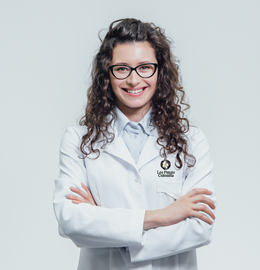 Médico sonriente con bata blanca y gafas, con los brazos cruzados sobre un fondo gris claro.