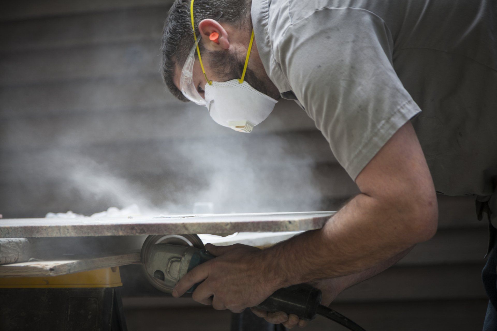 Uomo che usa una smerigliatrice elettrica, creando polvere. Indossa maschera e protezioni acustiche. Al chiuso, lavora sul legno.