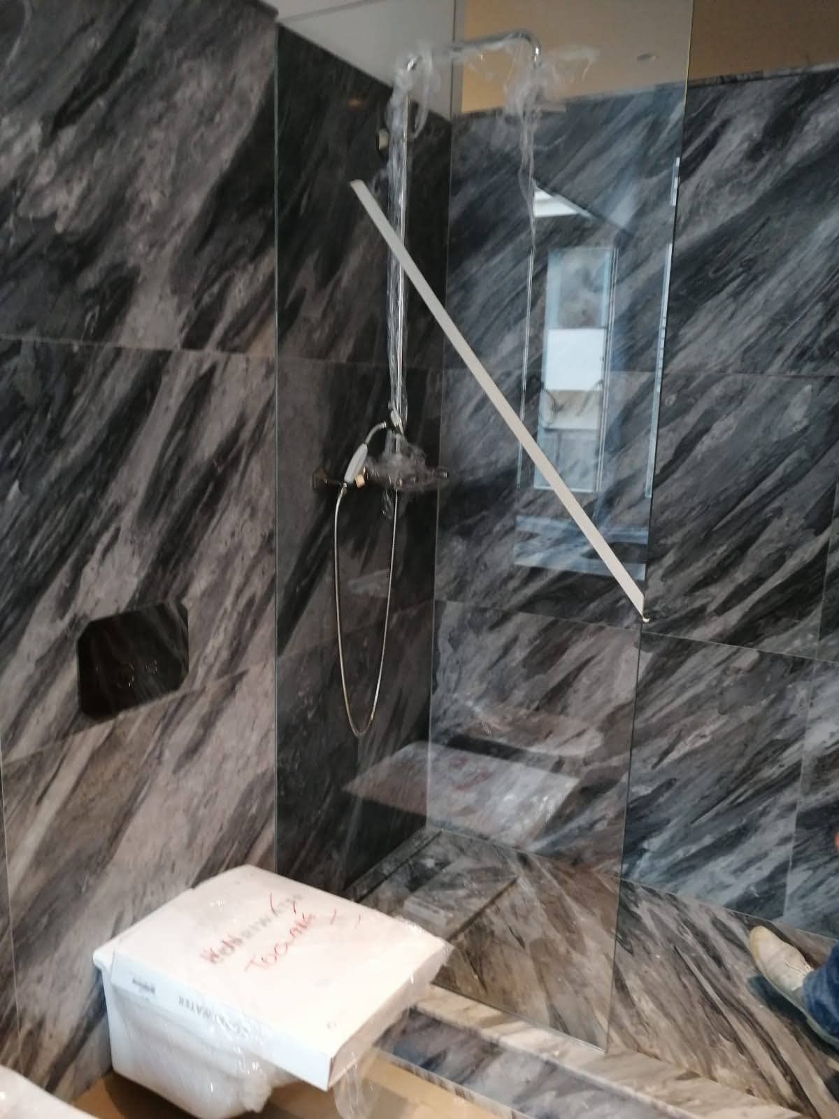Doccia del bagno con piastrelle in marmo grigio e bianco, soffione in vetro trasparente e un oggetto bianco in basso.