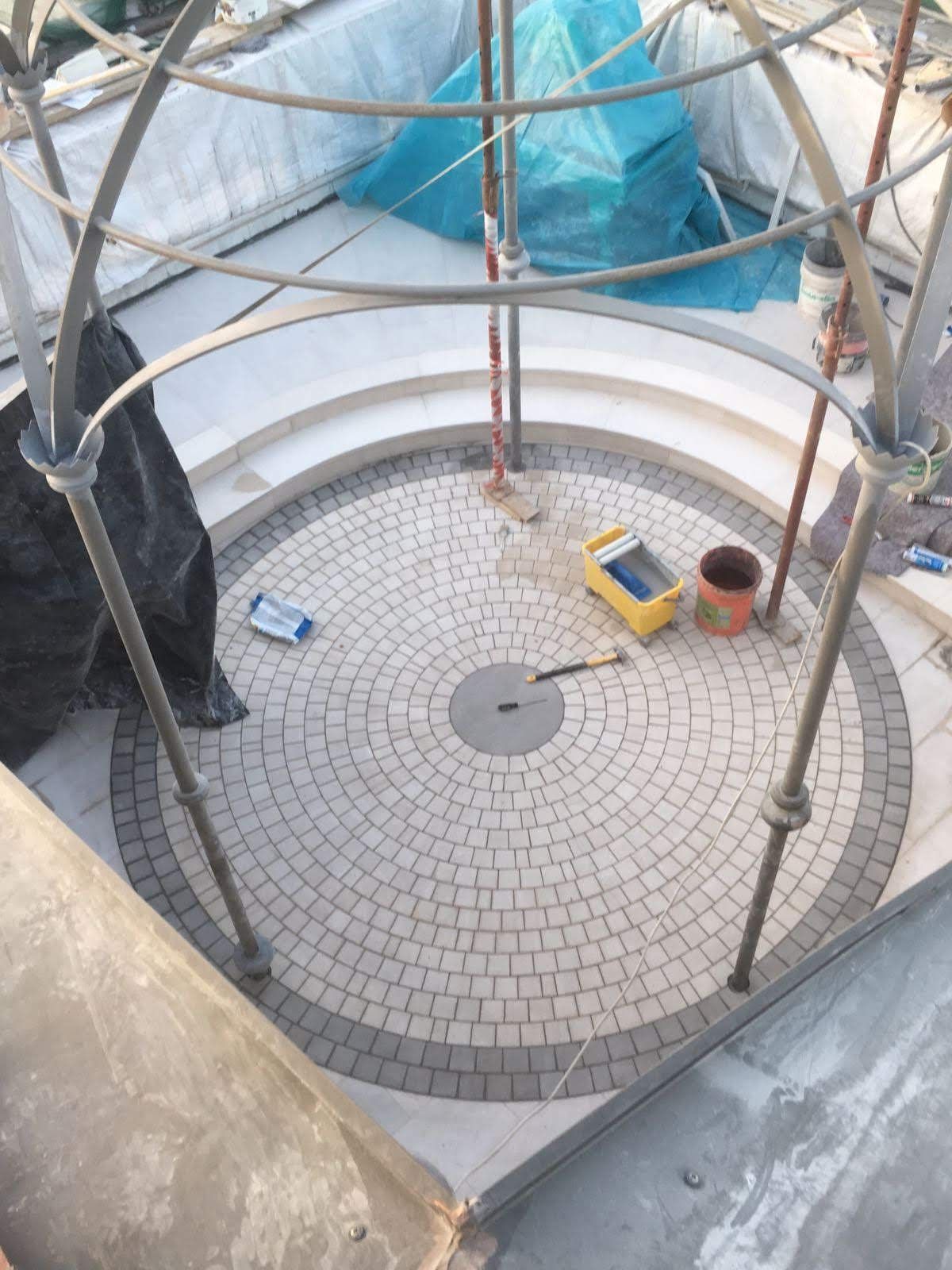 Patio circolare pavimentato con motivo radiale di ciottoli, struttura metallica e attrezzi da costruzione.