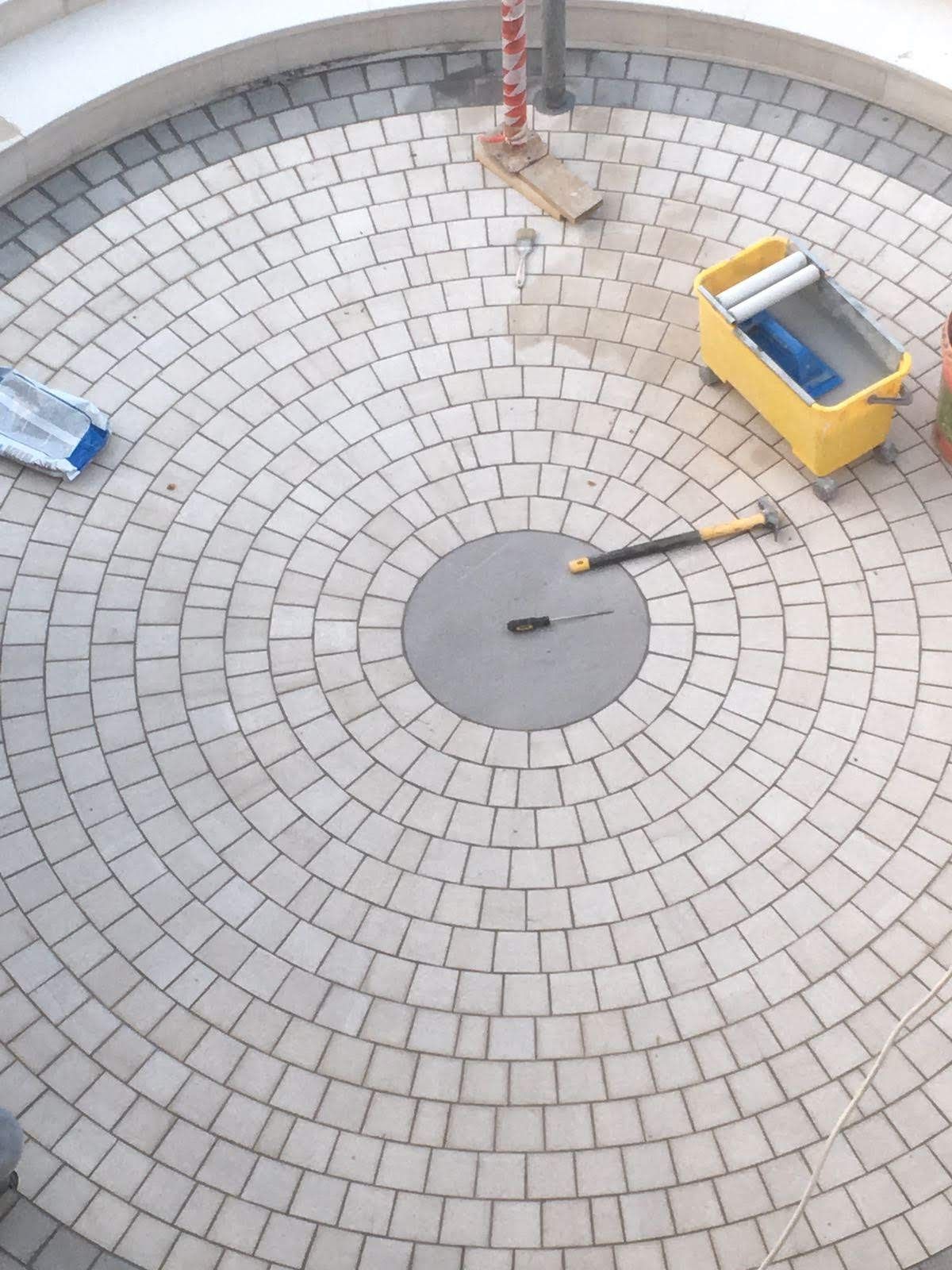 Area circolare pavimentata in costruzione con pietre grigie e chiare. Attrezzi e materiali sono sparsi tutt'intorno.
