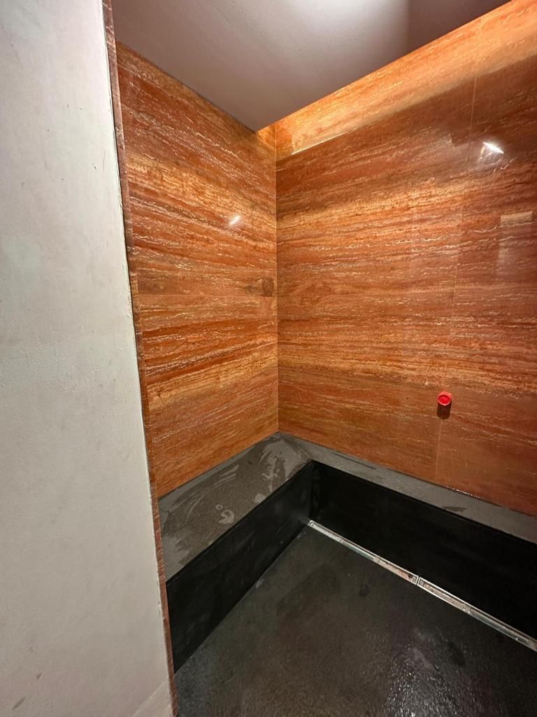 Angolo di una cabina doccia con pareti piastrellate di colore rosso-marrone, pavimento nero e una parete bianca parzialmente visibile.