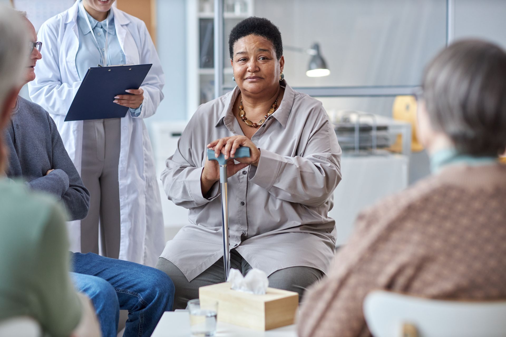 Small support group meeting with a speaker holding a cane and a doctor taking notes in a bright room