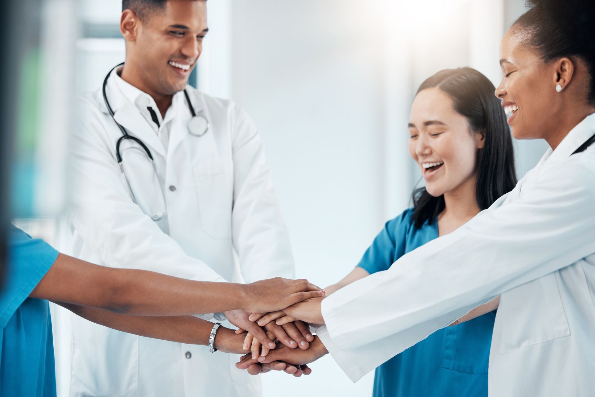 Healthcare team stacking hands in a hospital hallway, smiling in white coats and blue scrubs