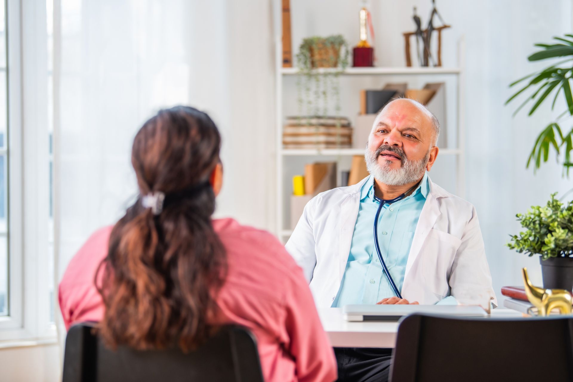 Doctor speaking to a patient in a bright office, seated at a desk with a laptop and plants nearby.