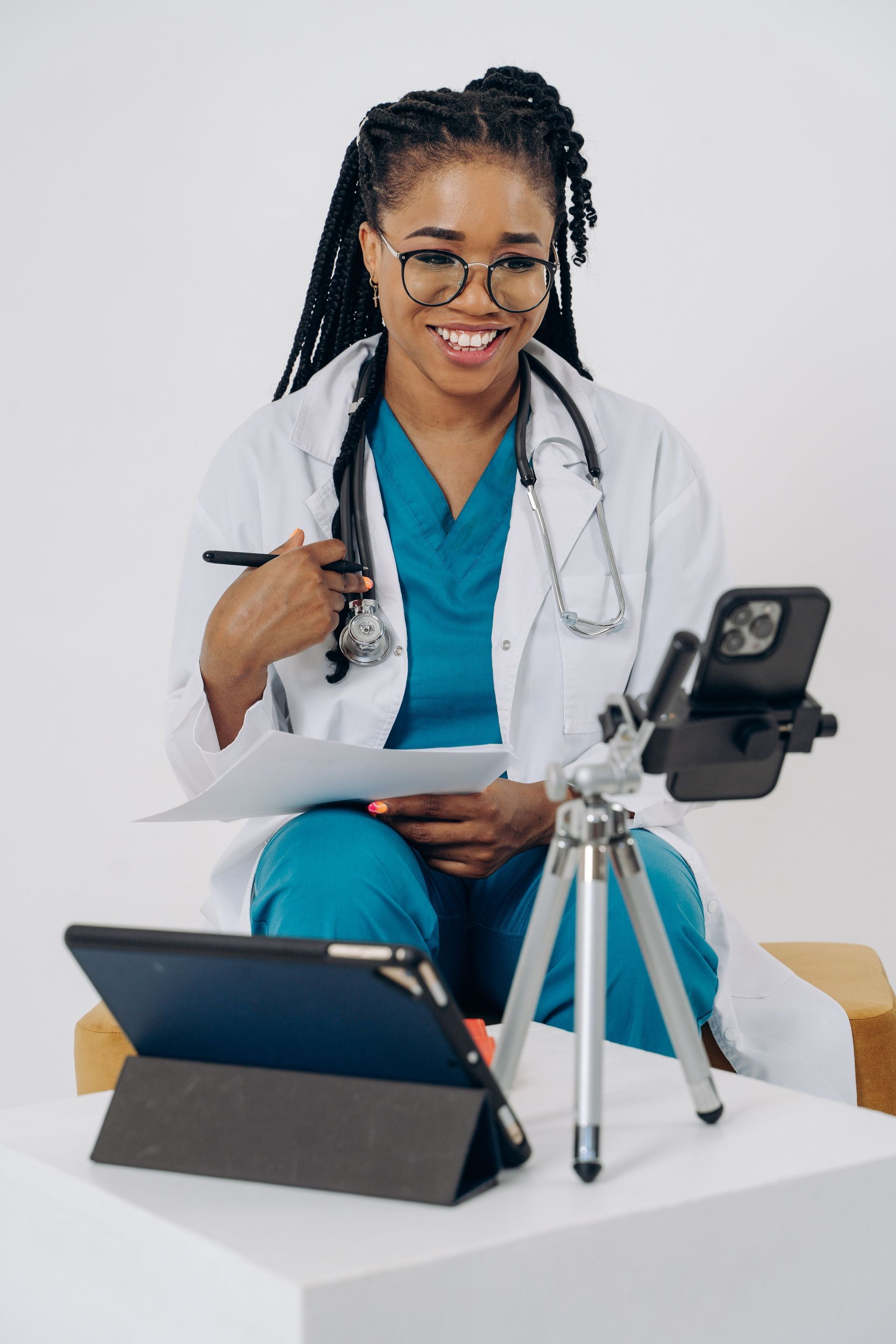 Doctor speaking during a telehealth video call, with a tablet on a tripod and laptop on the table.