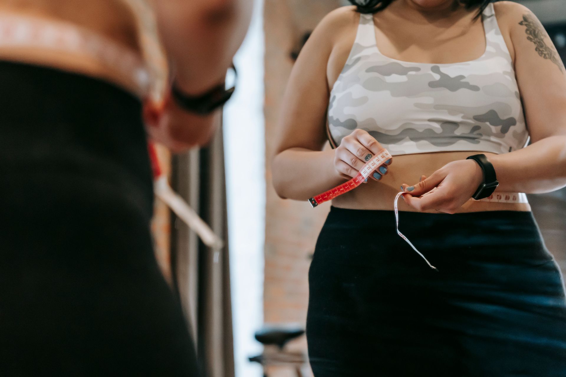 Person holding a red measuring tape in a gym, wearing a gray camo sports bra and black leggings