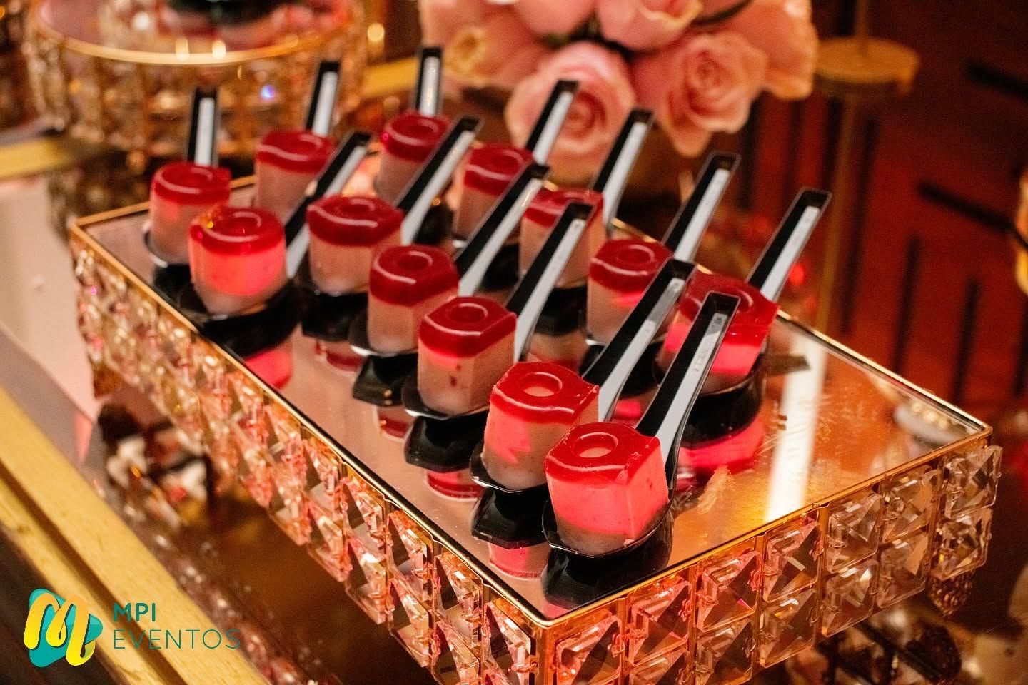 Vasitos de postre rojos y blancos con cucharillas negras dispuestos sobre una bandeja espejada en un evento formal