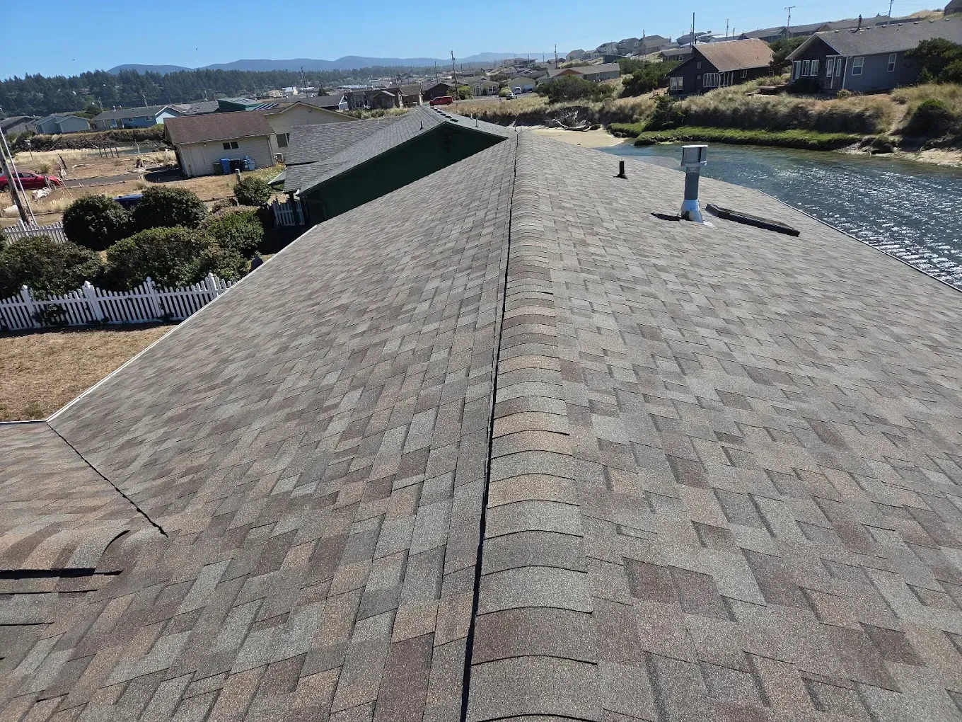View of a roof covered with brown and grey shingles, with a chimney.  Houses and water in the background.