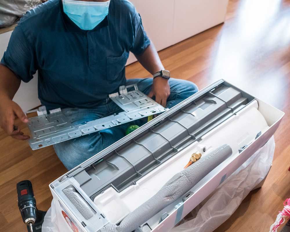 Person in scrubs kneels beside an open air conditioner unit, measuring parts with a ruler in a room.