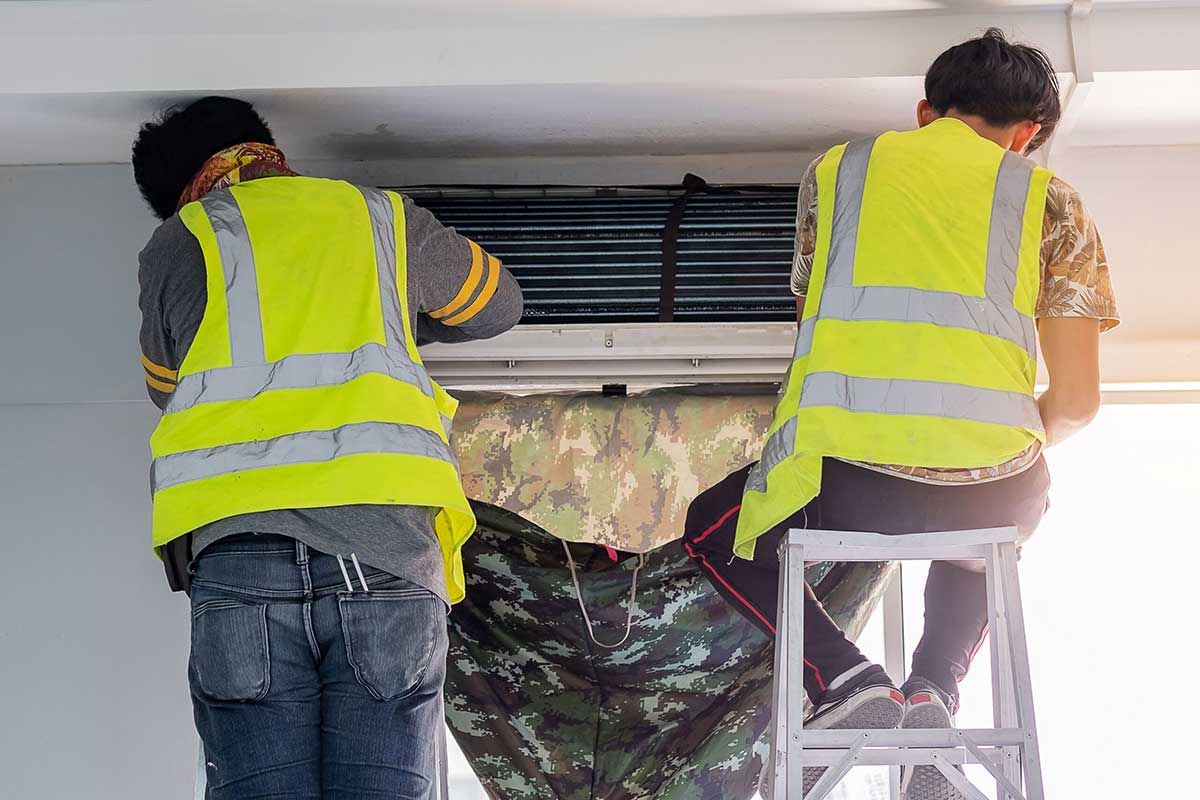 Two workers in yellow safety vests installing an air conditioner indoors using a ladder
