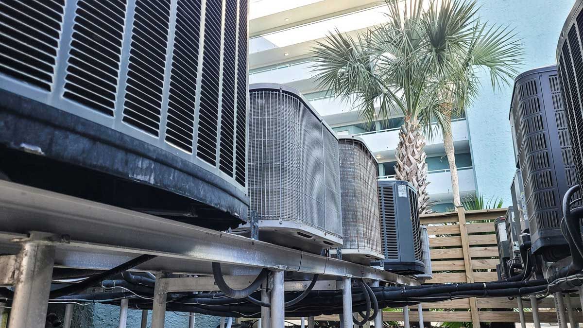 Row of large outdoor air conditioning units beside palm trees and a wooden deck under a canopy.