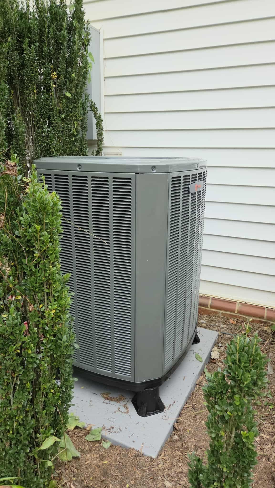 Outdoor HVAC unit beside a house wall, partially hidden by shrubs, on a concrete pad.