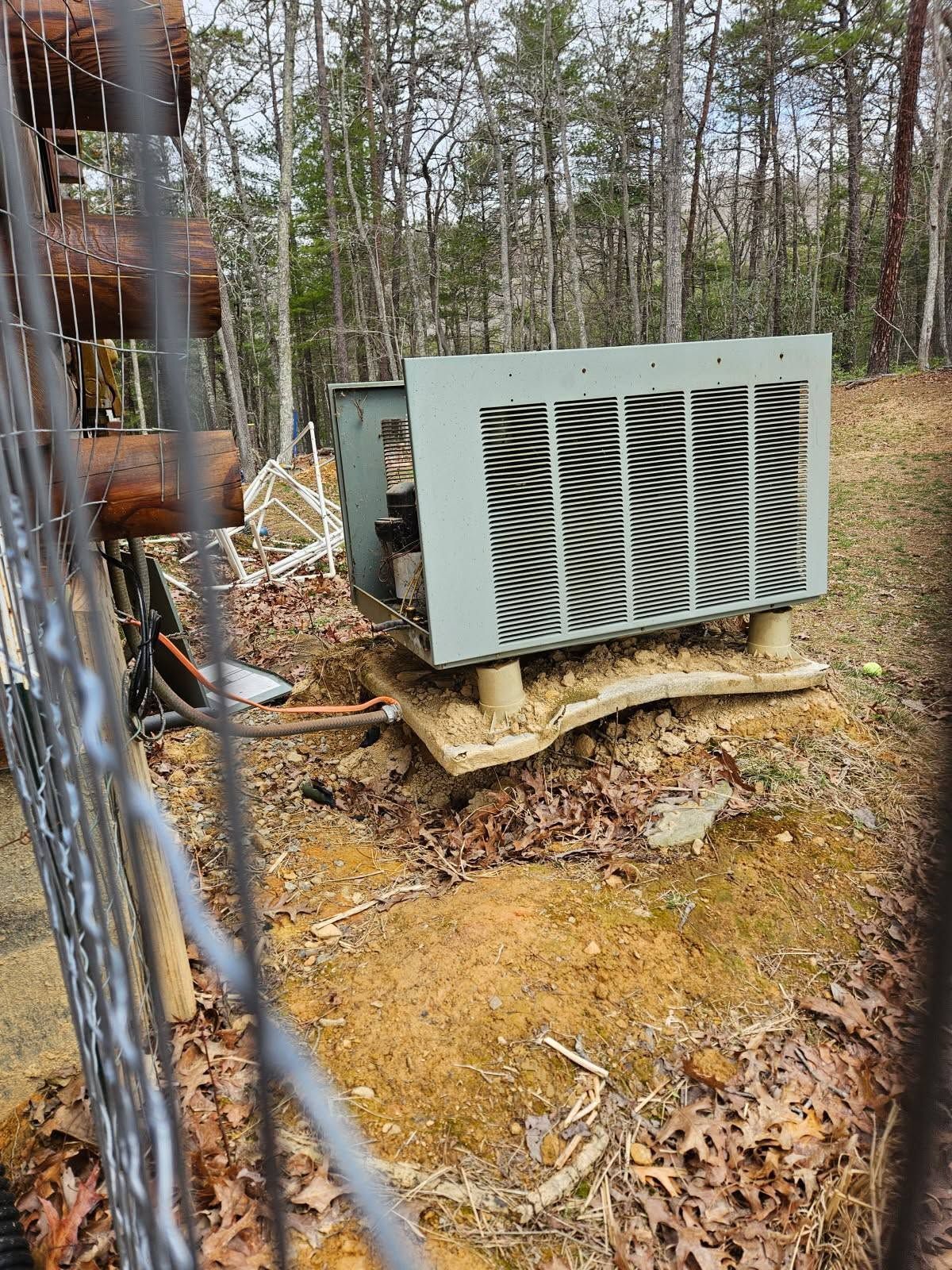 Outdoor HVAC unit on a concrete pad beside a chain-link fence in a wooded area