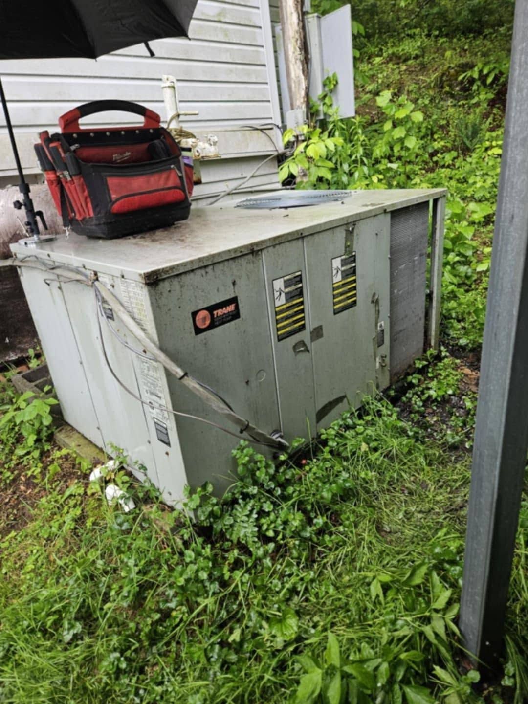 Gray outdoor HVAC unit beside a house, with a red tool bag on top and overgrown weeds around it