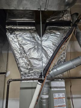 Foil-wrapped HVAC ductwork in a utility room with pipes and a small window below.