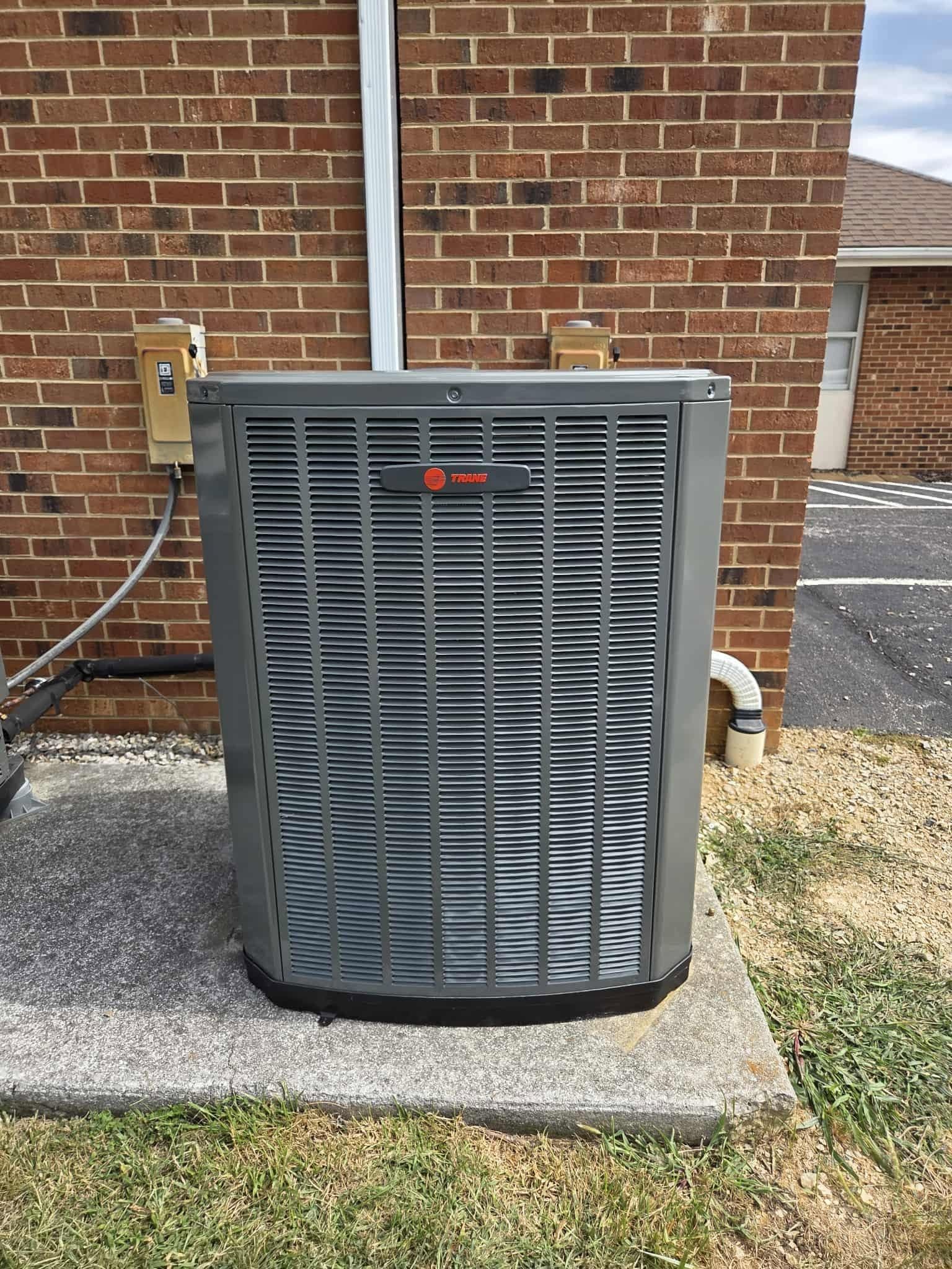 Outdoor air conditioner unit beside a brick wall, with gray grille and copper pipes.