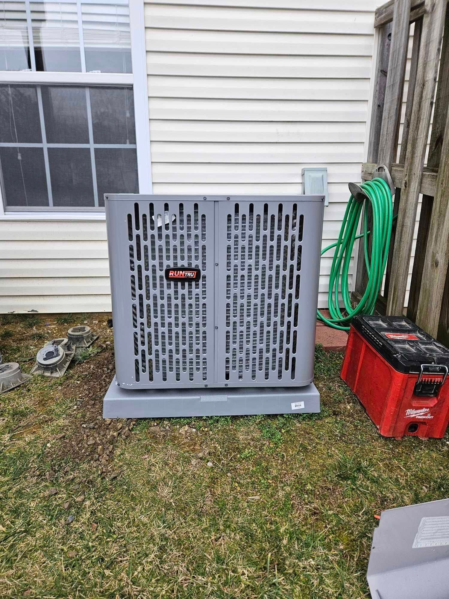 Gray vented equipment panel on lawn beside house, with red tool box and green hose nearby.