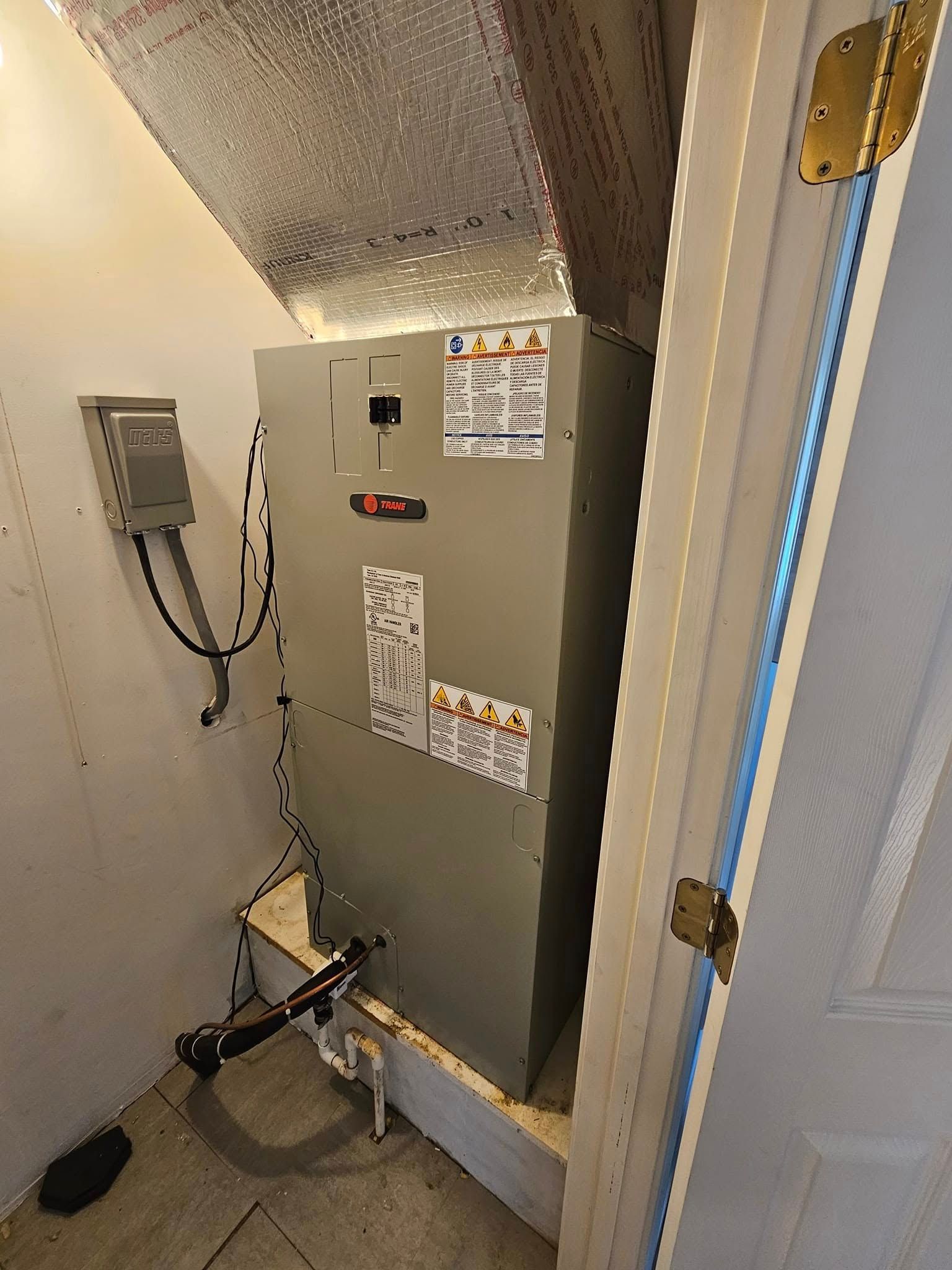 Gray HVAC unit in a narrow utility closet with warning labels and wiring.