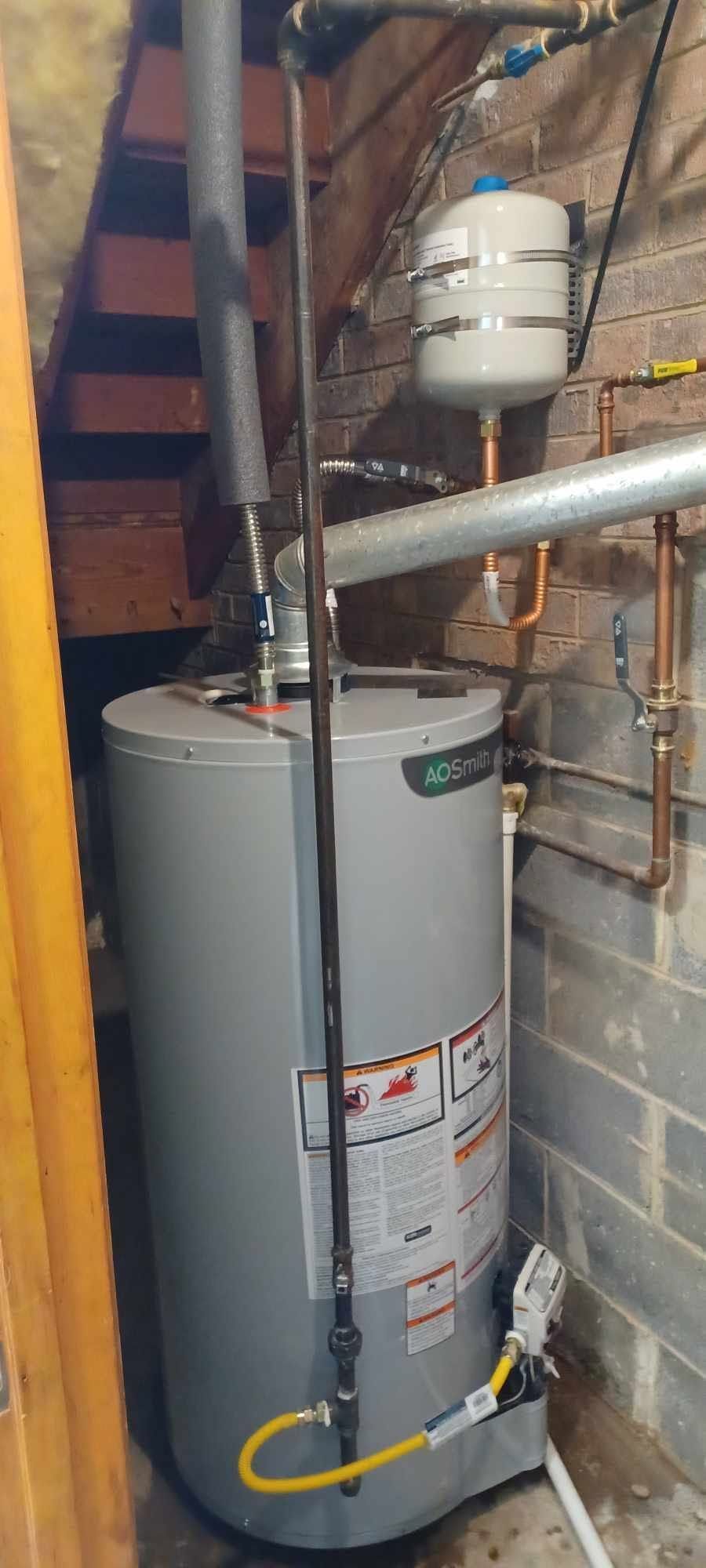 Water heater in a basement beside exposed pipes and a utility meter on a brick wall