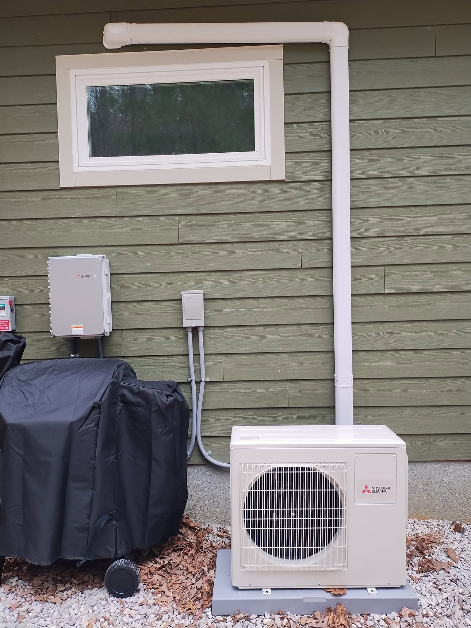 Outdoor HVAC unit and ductwork mounted on a green house wall beside a covered grill and utility boxes
